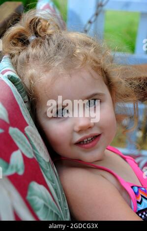 Das kleine Mädchen genießt es, auf der Veranda zu schwingen. Sie schaut seitwärts mit dem Kopf auf dem Schaukelkissen. Locken sind ihrem Pferdeschwanz entkommen. Stockfoto