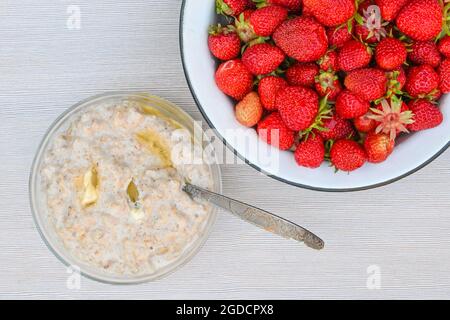 Frische reife, leckere Erdbeeren in einer Schüssel aus weißem Metall und ein Teller Haferbrei mit Butter auf einem weißen Tisch. Stockfoto