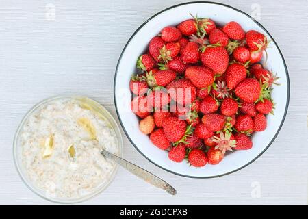 Frische reife, leckere Erdbeeren in einer Schüssel aus weißem Metall und ein Teller Haferbrei mit Butter auf einem weißen Tisch. Stockfoto