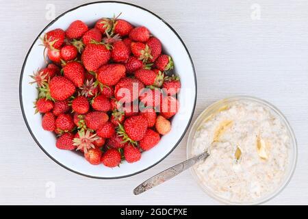 Frische reife, leckere Erdbeeren in einer Schüssel aus weißem Metall und ein Teller Haferbrei mit Butter auf einem weißen Tisch. Stockfoto