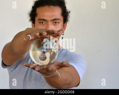 Ein hübscher junger Mann mit schmutzigen Fingernägeln hält in seinen Händen eine Kristallkugel mit seinem eigenen vertikal gespiegelten Spiegelbild und schaut auf den Betrachter. Stockfoto