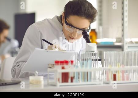 Nahaufnahme Wissenschaftlerin, die im Labor mit Mikroskop und Klemmbrett arbeitet Stockfoto