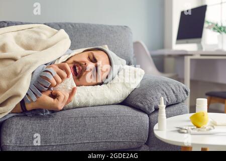 Junger Mann, der an Grippe oder Erkältung leidet, liegt auf dem Sofa und niest in Papiergewebe Stockfoto