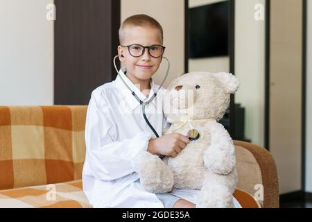 Niedliche kleine kaukasischen Jungen spielen lustige Spiel wie Arzt hält Stethoskop hören Spielzeug Vorschulkinder vorgeben, eine Krankenschwester zu sein behandeln flauschigen Patienten zu Hause. In einem Morgenmantel und einer medizinischen Maske. Stockfoto