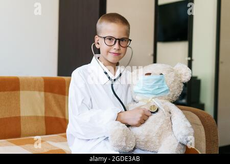 Blick auf ein Kind, das zu Hause im Sonnenlicht einen Arzt oder eine Krankenschwester mit einem Teddybären spielt. Happy Boy hört einem Spielzeug Stethoskop zu. Rollenspiel eines verspielten Kerls. In einer Brille und einem Gewand mit Schutzmaske Stockfoto