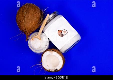 Kokosöl in einem Glas mit einem Holzlöffel und frischen Kokosnüssen, auf blauem Hintergrund Stockfoto