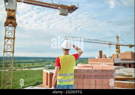 Mann in einem Hardhut, der vor Kranichen stand und rote Ziegel stapelte Stockfoto