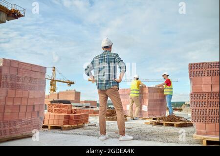 Vorarbeiter beobachten Arbeiter, die sich auf den Stapel roter Ziegelsteine stützen Stockfoto