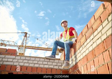 Nette ruhige Baumeister in einem Hardhut auf einer Ziegelstruktur sitzen Stockfoto