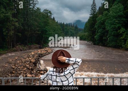 Frau in braunem Hut, die nach dem Sturm im Sommer auf den Gebirgsfluss schaut Stockfoto