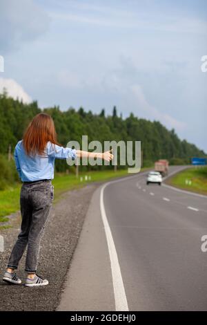 Das Mädchen hält das Auto mit der Hand auf der Autobahn an. Stilvolle Frau auf der Straße hält das Auto auf eine Reise gehen. Eine Straße mitten im Wald. Stockfoto