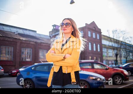 Attraktive Frau in Sonnenbrillen auf dem Hintergrund eines alten Backsteingebäudes. Eine Frau in der Stadt in einer leuchtend gelben Jacke vor dem Hintergrund eines Parkplatzes. Stockfoto