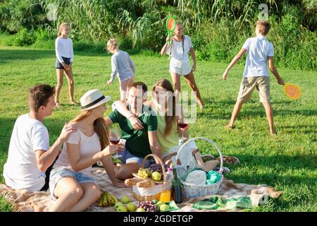 Frauen und Männer picknicken im Sommerpark mit glücklichen Kindern, die dahinter spielen Stockfoto