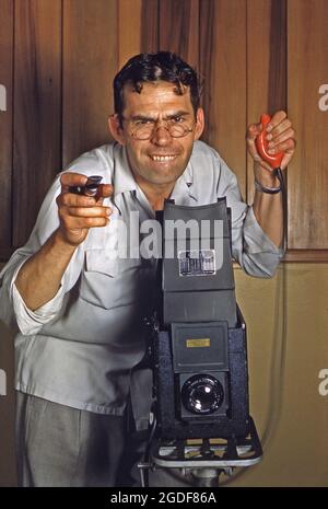 Studiofotografie in Amerika in den 1950er Jahren – ein Fotograf konzentriert sich hart und drückt mit seiner riesigen Graflex-Kamera einen Auslöserauslöser für die Glühbirne. Die Lampe (ein abnehmbares pneumatisches Gummigerät an einem Kabel, das mit der Kamera verbunden ist) bedeutete, dass der Bediener Verwacklungen der Kamera vermeiden konnte. Außerdem war es sehr nützlich, die Lampe gedrückt zu halten und das Objektiv offen zu halten, um Langzeitbelichtungen zu ermöglichen. Graflex war ein amerikanischer Kamerahersteller. Dieses Bild stammt aus einer alten amerikanischen Kodak Amateur-Farbtransparenz – einem Vintage-Foto aus den 1950er Jahren. Stockfoto