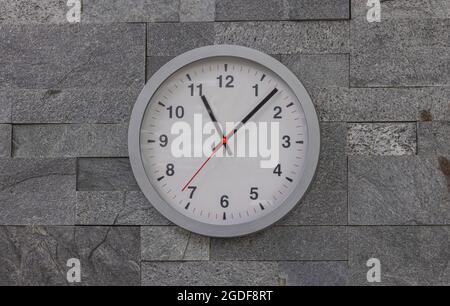 Moderne Wanduhr an einer Wand mit rustikalen Fliesen gefliest. Moderne weiße Wanduhr im Sonnenlicht mit Schatten auf dem Zifferblatt auf grauem Hintergrund. Nahaufnahme Stockfoto