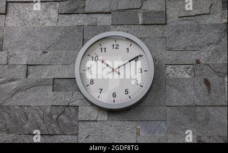 Moderne Wanduhr an einer Wand mit rustikalen Fliesen gefliest. Moderne weiße Wanduhr im Sonnenlicht mit Schatten auf dem Zifferblatt auf grauem Hintergrund. Nahaufnahme Stockfoto