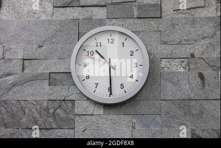 Moderne Wanduhr an einer Wand mit rustikalen Fliesen gefliest. Moderne weiße Wanduhr im Sonnenlicht mit Schatten auf dem Zifferblatt auf grauem Hintergrund. Nahaufnahme Stockfoto