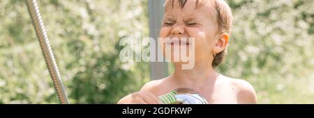 Der kleine vierjährige Squint-Kid-Boy Fun gießt sich im Sommer bei der Hitze in der Natur aus einer Gießkanne aus einer Außendusche mit kaltem Wasser Stockfoto