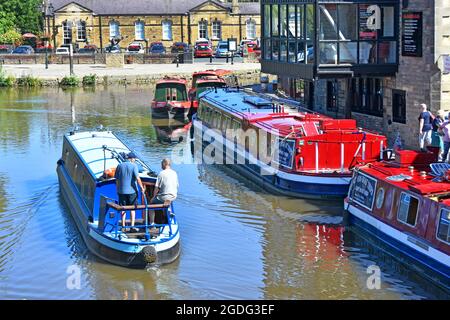 Rückansicht von zwei Personen auf einem schmalen Boot, die Leeds zum Liverpool-Kanal bei Skipton fahren und dabei an festgetäuten Passagierrundfahrten durch North Yorkshire England, Großbritannien, vorbeifahren Stockfoto