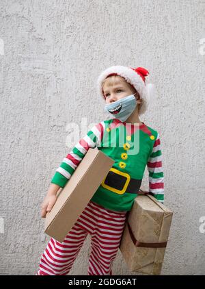 Kleinkind Junge gekleidet als Weihnachtself, Santa Helfer. Das Tragen einer blauen medizinischen Maske mit einem Lächeln auf der Maske. Er hält Kisten in den Händen. Positiv festlich bei Stockfoto