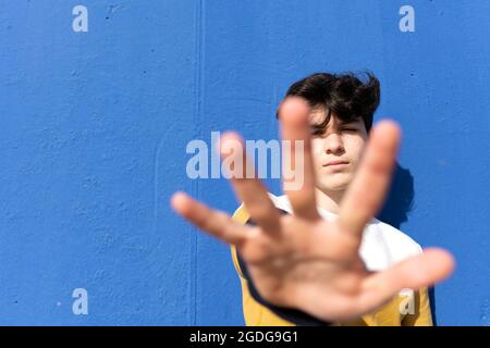 Junger Mann, der sich an einer blauen Wand lehnte und sein Gesicht unter verschwommener Hand versteckte Stockfoto