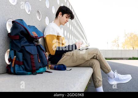 Junger Mann schreibt Notizen in einem Notizbuch, das auf einer Bank sitzt Stockfoto