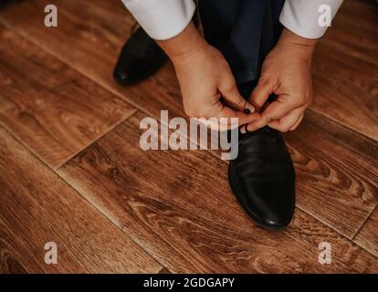 Ein Mann in einem weißen Hemd und einer dunklen Hose sitzt auf einem Stuhl und bindet die Schnürsenkel seiner schwarzen Stiefel auf einem braunen, strukturierten, glänzenden Holzboden. Herren Stockfoto