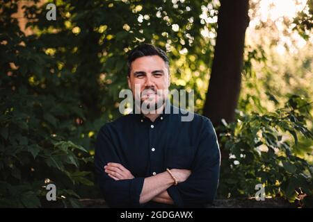 Porträt eines glücklichen Geschäftsmannes mit gekreuzten Armen, der im Sommer gegen Bäume steht Stockfoto