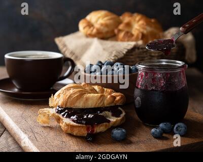 Croissant mit Heidelbeermarmelade und Heidelbeeren auf einem Holzbrett mit Kaffee, einer Schüssel mit Beeren und einem Korb mit Croissants im Hintergrund Stockfoto