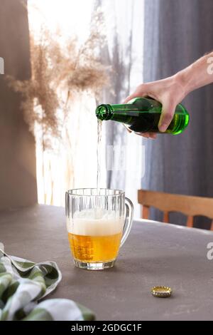 Ein Mann gießt leichtes Bier in ein Glas aus einer Flasche, ein Glas Bier mit Schaum auf einem grauen Tisch vor dem Hintergrund eines Fensters Stockfoto