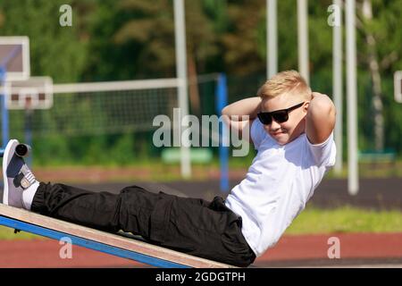 Ein junger Mann in dunkler Brille trainiert im Sommer auf dem Sportplatz die Muskeln der Presse. Hochwertige Fotos Stockfoto