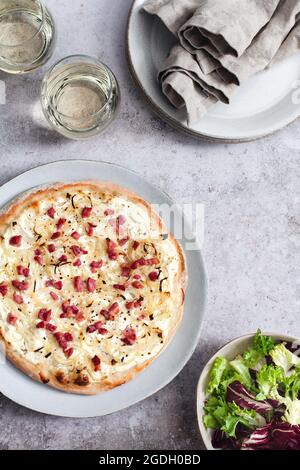 Flammkuchen mit Creme fraiche, Lardonen und Zwiebeln, serviert mit einer Schüssel Beilagensalat und zwei Gläsern Weißwein. Stockfoto