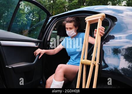 Eine Frau mit einem verletzten Bein steigt in ein Auto. Orthopädischer Putz, orthopädische Krücken. Eingeschränkte Mobilität Stockfoto