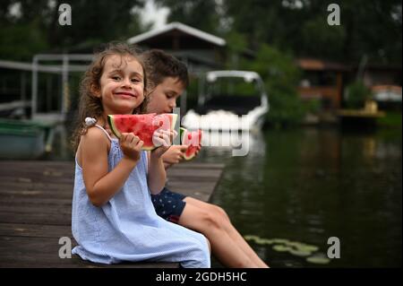 Hübsches kleines Mädchen in einem Kleid, das sich auf dem Pier neben ihrem Bruder ausruht, eine Wassermelone in den Händen hält und sanft lächelt, während sie die letzte Wärme genießt Stockfoto