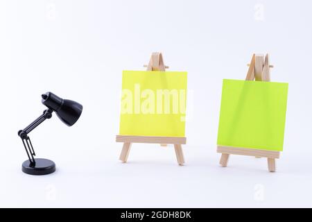 Zwei hölzerne Easels Miniature mit blanker quadratischer Leinwand oder Memopapier - Mockup. Mini-Holzständer mit sauberer Tafel und kleiner schwarzer Tischleuchte auf weißem Hintergrund, Kopierraum Stockfoto