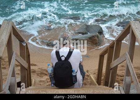Reifer Mann mit grauem Haar und einem Rucksack, der auf einer Holzleiter am Meer sitzt Stockfoto
