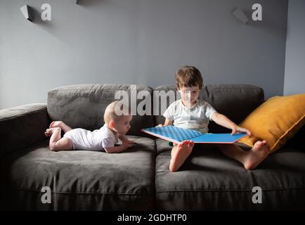 Der Junge liest ein Buch, das auf dem Sofa sitzt und sein Bruder liegt in der Nähe Stockfoto