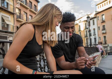 Junger schwarzer Junge und blondes kaukasisches Mädchen, das auf einem Stadtplatz sitzt. Er zeigt ihr etwas auf seinem Handy. Stockfoto