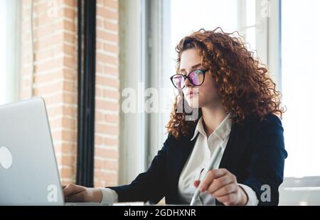 Schöne junge lockige rothaarige Frau in einem Business-Anzug und Die Brille funktioniert im Büro auf einem Laptop Stockfoto