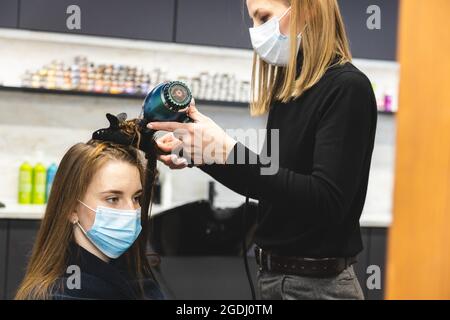 Meisterin Friseur in einer medizinischen Maske trocknet das Haar des Mädchens mit einem Haartrockner und Kämme nach dem Waschen in einem Schönheitssalon. Covid-19 Pandemie und Stockfoto