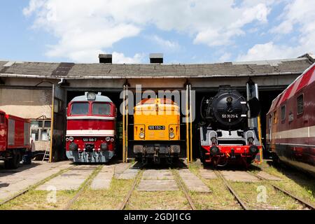 Elektrische Triebwagen und eine Dampflokomotive stehen in einem Lokschuppen vor blauem Himmel und Wolken. Stockfoto