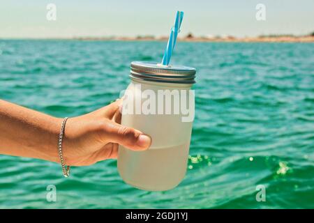 Kleine Flasche mit einem Stroh in einer weiblichen Hand auf einem Hintergrund des Meeres Stockfoto