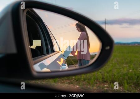 Rückspiegel mit einer Spiegelung der weiblichen Silhouette lädt ein Elektroauto, im Hintergrund sichtbaren Himmel bei Sonnenuntergang Stockfoto