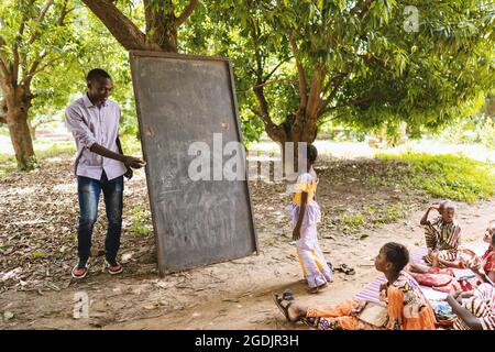 Großer schwarzer Lehrer in legerer Kleidung lädt einen seiner kleinen Schüler ein, in einer Freilichtschule in Westafrika etwas auf die Tafel zu schreiben Stockfoto