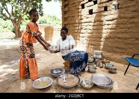 Schwarzer afrikanischer Koch sitzt auf einem kleinen Holzstuhl in der Mitte von Tellern und Küchenutensilien und bietet einem Mädchen, das neben ihm steht, eine Schüssel Reis an Stockfoto