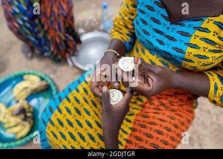 Fürsorgliche Afrikanerin in einem bunten Kleid sitzt auf dem Boden in einer ländlichen Dorfküche und teilt sich ein halbes Ei mit einem Kind; Armutskonzept Stockfoto