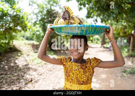 Auf diesem Bild steht ein hübsches schwarzes Mädchen auf einer Landstraße in Westafrika und trägt einen Teller voller Bananen auf dem Kopf Stockfoto