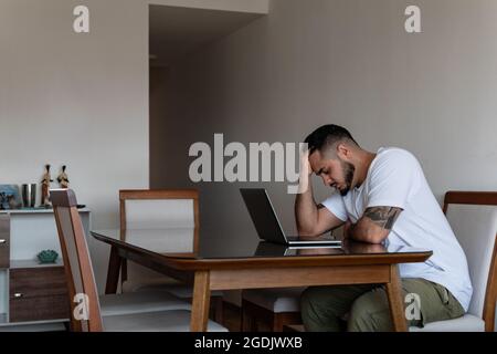 Der junge tätowierte lateinische Mann vor dem Laptop stresste heraus und hielt seinen Kopf fest Stockfoto