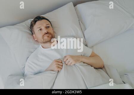 Einsamer verstörter junger bärtiger Mann, der mit offenen Augen im Bett liegt Stockfoto