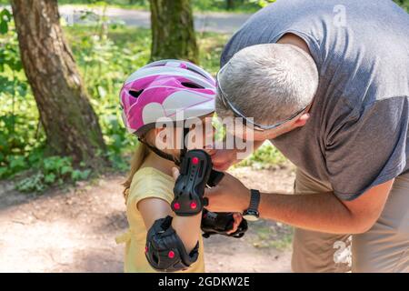 Vater-Vater setzt sich einen Schutzhelm für ein Fahrrad auf seine Tochter. Hilfe und Pflege für Kinder. Sicherheit in der Stadt auf einem Roller oder Fahrrad Stockfoto